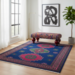 A colorful area rug with a deep blue base and geometric motifs in pink, green, and red, styled in a bright room with a matching patterned bench, a gold floor lamp, a potted plant, and a framed abstract print.