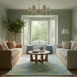 A beautifully styled living room featuring a large abstract-patterned area rug in sage green, light blue, and cream, flanked by two beige sofas and a round wooden coffee table. The room is decorated with a potted fiddle-leaf fig, a gold chandelier, and a bay window looking out onto a lush green garden.