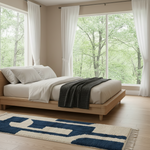 A minimalist light wood platform bed dressed in white bedding and a dark textured throw blanket, set in a bright bedroom with large windows looking out onto a green forest. A blue and cream geometric patterned rug is on the floor in front of the bed.