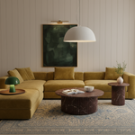 A modern living room featuring a large mustard yellow sectional sofa with matching pillows, styled with a round marble coffee table, a side table, a green table lamp, and a patterned rug against a beige paneled wall.