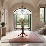 A large pink area rug with blue geometric diamond patterns is displayed in a bright, elegant entryway with a wooden pedestal table and vase in the center. The room features cream-colored walls, arched windows, a staircase, and a view of a green garden outside.