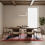 A dining room scene with a rectangular wooden dining table and four matching chairs arranged on a colorful striped rug beneath a white pendant light. The room has beige walls, exposed wooden ceiling beams, a large bright window, and a potted plant.