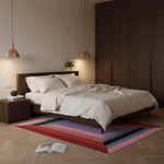 A modern bedroom scene with a bed dressed in white bedding, a dark wood headboard and nightstand, a large dark wood wardrobe, two pendant lights, a colorful striped rug, and an open book on a herringbone wood floor.