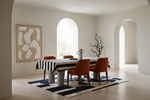 A styled dining room interior with a rectangular table covered in a black-and-white striped tablecloth, surrounded by four orange-brown upholstered chairs. A dark blue and white patterned rug lies beneath the table, and the room has warm off-white walls, arched windows, and abstract wall art.