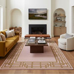 A styled living room featuring a large rectangular area rug with a beige geometric border and a dusty pink center, placed under a coffee table in front of a fireplace.