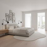 A minimalist bedroom featuring a low-profile platform bed with light beige upholstery, dressed in neutral cream bedding. The room has light wood floors, a matching wavy cream rug, black wall-mounted lamps, and a gallery wall of framed abstract art.