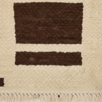 Close-up of a textured woven rug or textile in warm cream with two dark brown rectangular woven sections and a cream fringe along the bottom edge.