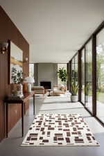 A modern living room interior with a rectangular area rug in front of large floor-to-ceiling windows. The rug has an off-white background with a geometric pattern of dark brown rectangles. The room features warm wood paneling, a console table, and indoor plants.