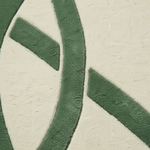 A close-up of a textured rug or carpet showing a dark green abstract geometric pattern on a warm off-white background.