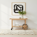 A light natural wood rolling side cart with black wheels stands on a cream rug in a bright room. A glass vase with greenery and a small ceramic object sit on the top shelf, and a framed abstract black-and-white print hangs on the wall above.