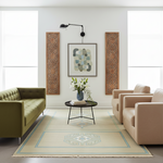 A styled living room scene featuring a large beige area rug with a blue geometric medallion pattern, flanked by a green tufted sofa and beige armchairs. The room is decorated with two vertical carved wood panels and a central framed abstract print.