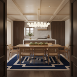 A modern dining room viewed through dark wood door frames, featuring a light wood dining table and six woven cane chairs on a bold blue and white geometric rug. The room has a coffered ceiling, a contemporary glass chandelier, and a kitchen in the background.