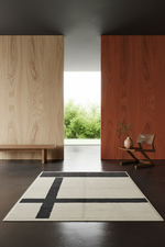 A modern area rug with a black geometric cross pattern on a cream background, styled in a minimalist, contemporary interior with wood-paneled walls and a dark floor.