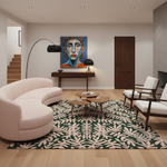 A modern living room featuring a curved blush pink sofa, a green and beige patterned area rug, two wooden coffee tables, and two white armchairs with dark wood frames. A colorful portrait painting hangs on the wall above a dark wood console table.