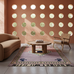 A styled living room scene featuring a rectangular area rug with a geometric medallion pattern in dusty pink, teal, and cream, placed on a light gray tiled floor. The room has a terracotta-colored wall with circular cutouts, a curved beige sofa on the left, and a wooden armchair on the right.
