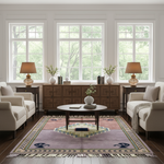 A styled living room with a large rectangular area rug featuring a geometric medallion pattern in muted purple, beige, and navy. The room is furnished with cream-colored armchairs, a wooden sideboard, two table lamps, and a round coffee table, all set against a bright background of three large windows showing green trees outside.