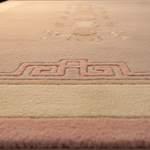 Close-up of a plush area rug with a soft pink border, a beige center, and a raised pink geometric pattern.