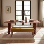 A styled interior scene showing a rectangular dining or study table with a pinkish-red marble top and matching marble legs, paired with a mustard-yellow upholstered bench. The table is set with books and a vase in a bright room with large black-framed windows, curtains, and a patterned rug.