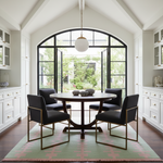 A modern dining room featuring a round dark wood table with four black upholstered chairs that have gold metal frames, set on a green and pink patterned rug in front of a large arched window.