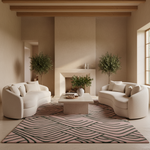 A styled living room featuring two curved cream boucle sofas facing a low concrete coffee table on a pink and black patterned area rug. The room has warm beige walls, a white fireplace, and two potted olive trees.