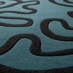 Close-up of a plush area rug with a teal blue background and thick, dark charcoal wavy lines forming an abstract pattern.