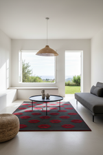 A modern living room featuring a dark gray rug with a repeating red lip pattern, styled with a round black coffee table, a woven pendant light, a gray sofa, and a round woven pouf, with large windows looking out to a green landscape.