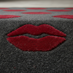 A close-up of a dark gray textured rug featuring a large red lips motif in the center and smaller red heart shapes along the top edge.