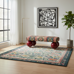 A large traditional-style area rug with a floral and geometric pattern in teal, beige, pink, and gold tones, styled in a bright modern room with a bench, floor lamp, and potted plant.