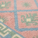 Close-up of a soft, textured rug with a pastel color palette. The design features pink squares outlined in light blue, decorated with light green botanical motifs and cream accents.