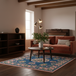 A styled living room scene featuring a rust-orange velvet sofa, a round dark wood coffee table, a blue and pink patterned area rug, and a white vase with a green plant in the center. The room has warm beige walls, dark wood ceiling beams, and a dark wood bookshelf.