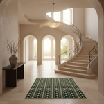 A rectangular area rug with a dark green and cream leaf pattern is displayed on a light beige tiled floor in a grand, sunlit entryway with a sweeping staircase and arched windows in the background.