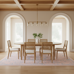 A modern dining room set featuring a round light oak wood table with woven cane seats, surrounded by six matching wooden chairs with cream upholstered seats. The set is styled on a pink patterned area rug under a brass multi-light chandelier, set against a warm beige wall with arched windows.