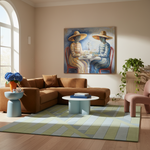 A large framed art print depicting two figures in striped clothing sitting at a table, hung on a beige wall in a modern living room with a brown sectional sofa, light blue coffee tables, and a striped green and blue rug.