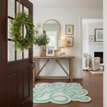 A styled entryway with a light green and white cloud-shaped rug on a dark wood floor, a wooden console table, a green wreath on a dark brown door, and a view into a living room.