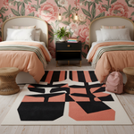 A modern geometric area rug with abstract black and coral-pink shapes on a cream background, styled in a bedroom setting between two beds with peach bedding and floral wallpaper.
