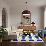 A modern living room featuring a cream sectional sofa, a blue accent chair, a round green coffee table, and a cream rug with large blue polka dots. A wooden sideboard with a large framed art print stands in the background under a dark red pendant light.