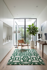 A modern living room scene featuring a prominent green and cream geometric patterned area rug on light wood flooring. The room is styled with a brown leather armchair, a large potted fiddle-leaf fig, a dark wood console table, and white cabinetry along the left wall.