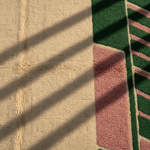 A close-up of a textured rug with a cream-colored center, a pink geometric panel, and green striped borders, cast with diagonal shadows from window blinds.
