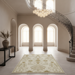 A rectangular area rug with an ornate beige and gold botanical pattern lies on a light stone floor in a grand, sunlit interior hallway with arched windows, a staircase, and a chandelier.