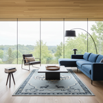 A styled modern living room with a large blue sectional sofa, a matching armchair, a low rectangular coffee table, a patterned area rug, a black floor lamp, and a small side table with books, all set on a light wood floor in front of floor-to-ceiling windows overlooking a forest.
