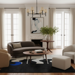 A styled modern living room featuring a dark brown curved sofa, a round wooden coffee table, a black and white abstract patterned rug, a white ottoman, and cream armchairs, set against large windows with beige curtains.