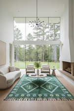 Modern living room with a green and white patterned rug, white furniture, and large windows overlooking trees.