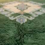 Close-up of a plush green rug with a textured shag pile and a blurred traditional pattern in cream, light blue, and beige in the background.