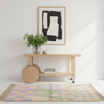 A light natural wood rolling console table with a lower shelf and caster wheels, styled in a bright room with a potted green plant, a small metallic sphere, and a framed abstract print on a white wall above. A multicolor patchwork area rug with fringe is spread on the floor in front of the table.