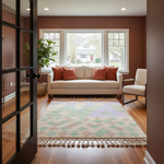 A styled living room interior featuring a cream sofa with rust-orange pillows, a multicolor geometric patterned area rug with fringe, a white armchair, a potted plant, and a black-framed glass door, set against warm brown walls.