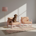Two blush pink armchairs with pale peach cushions are arranged in a sunlit room beside a pink floor lamp and a patterned rug. A small brown dog sits on the left chair.