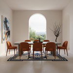 A modern dining room scene with a long oval table and eight orange upholstered chairs arranged on a geometric blue, white, and black rug. The room has warm off-white walls, a large arched window looking out to greenery, and decorative wall art and a black tree sculpture.