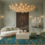 A luxury living room featuring a large, organic-shaped woven pendant chandelier with warm glowing lights. The room has elegant pale sage green walls with classical molding, a cream-colored curved sofa, a marble coffee table, a teal and gold patterned rug, and a green patterned armchair.
