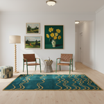 A styled interior room with a large teal area rug featuring a wavy gold pattern on the light wood floor. The room includes two green-framed chairs, a wooden floor lamp, a small stone side table, a patterned pouf, and three framed landscape and floral paintings on the white wall.
