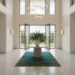 A modern interior entryway with a round stone pedestal table centered on a teal and gold patterned area rug. A large white vase with green foliage sits on the table. The room has tall windows, open black-framed doors, wall sconces, and a multi-ring chandelier overhead.